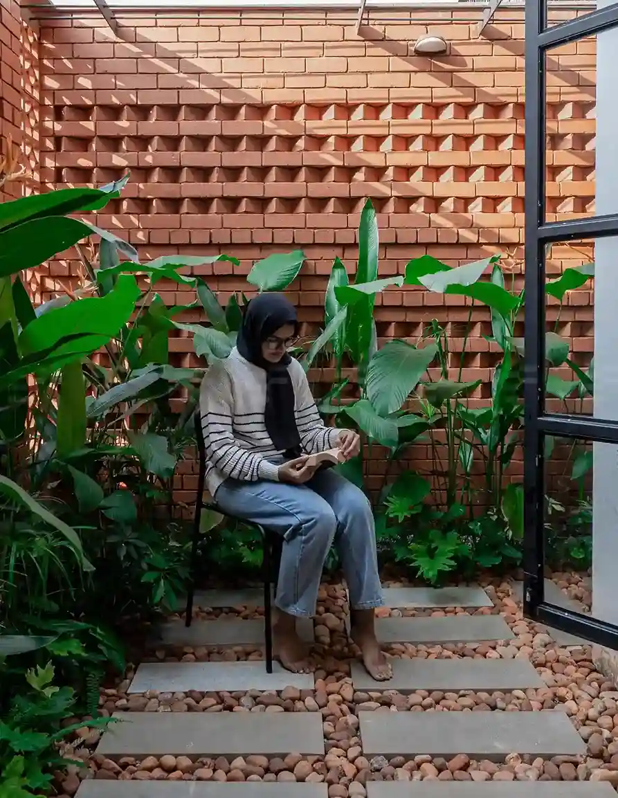 Cozy courtyard with a girl reading, surrounded by lush plants, brick walls, and stepping stones, designed by Form Architects in Malappuram.