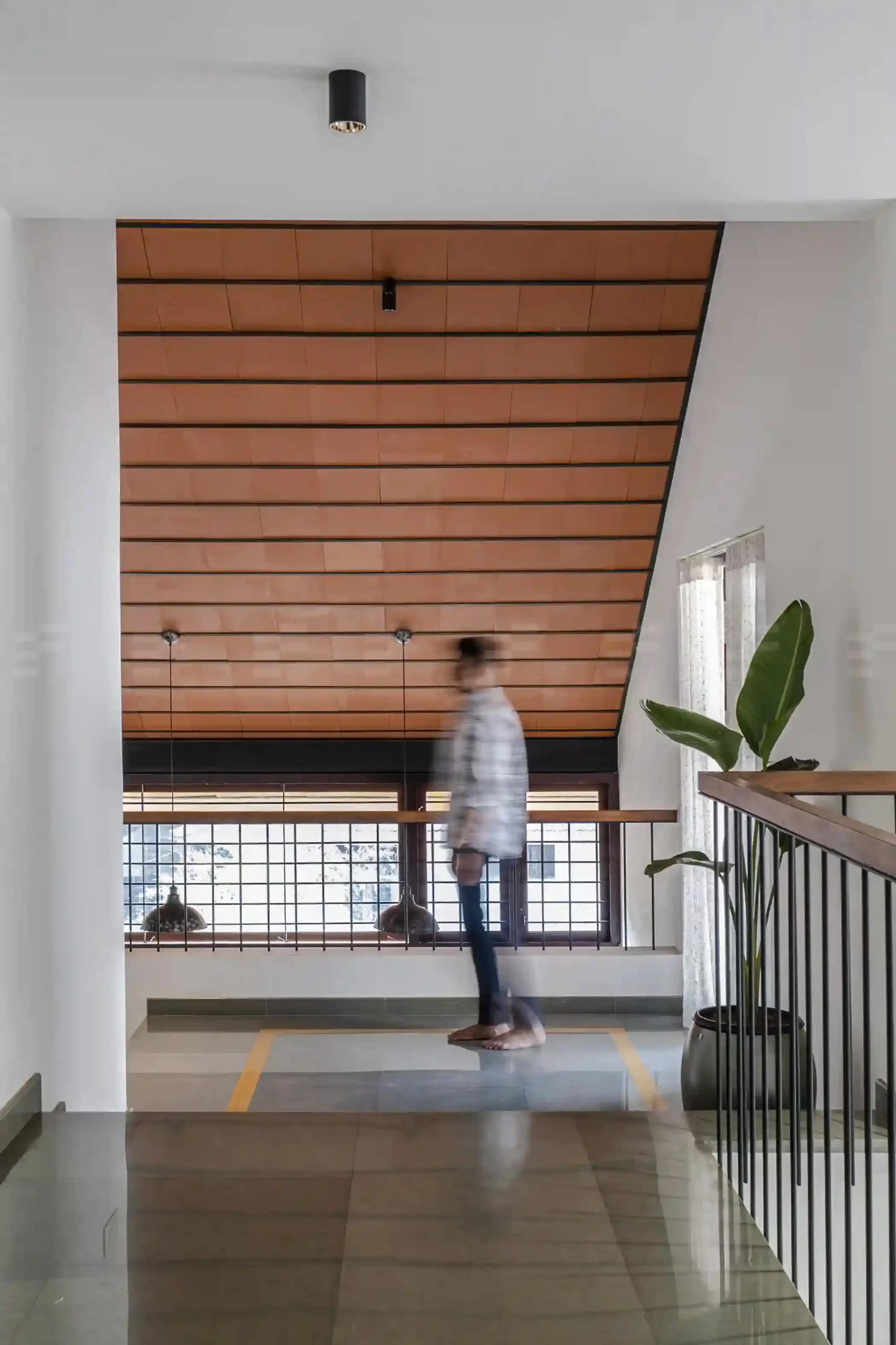 Upper floor landing in a Malappuram home with terracotta-tiled sloped ceiling, minimalist railing, and potted indoor plant.