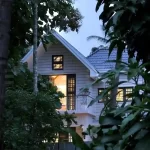 Evening view of a Kerala colonial style home with gabled roofs, white exterior walls, and surrounded by tropical plants.