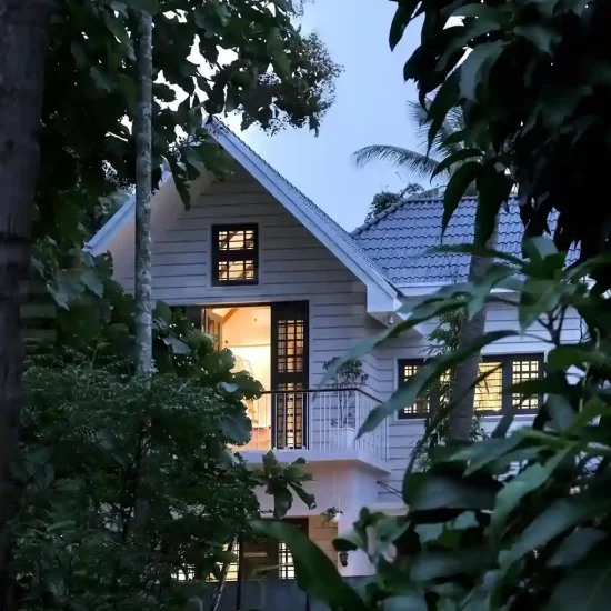 Evening view of a Kerala colonial style home with gabled roofs, white exterior walls, and surrounded by tropical plants.