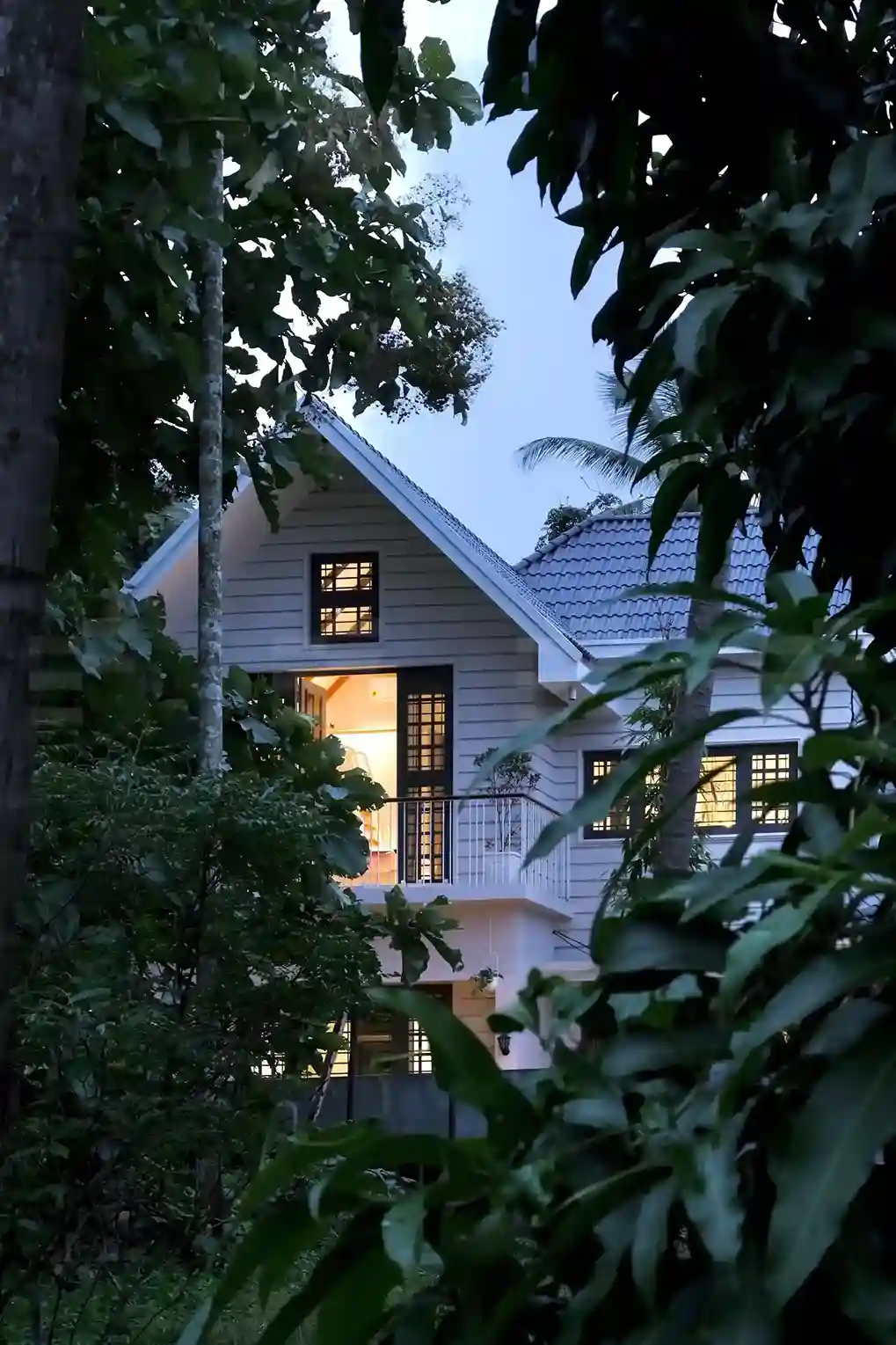 Evening view of a Kerala colonial style home with gabled roofs, white exterior walls, and surrounded by tropical plants.