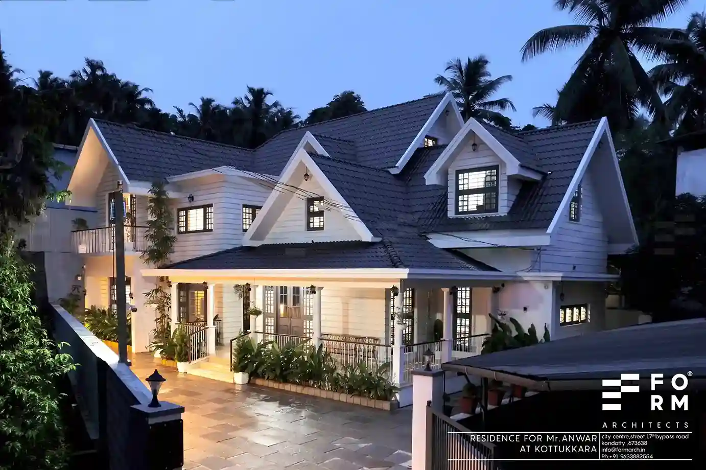 Grand entrance of Kerala colonial house design at night, with lit-up archways, double wooden doors, and lush greenery.