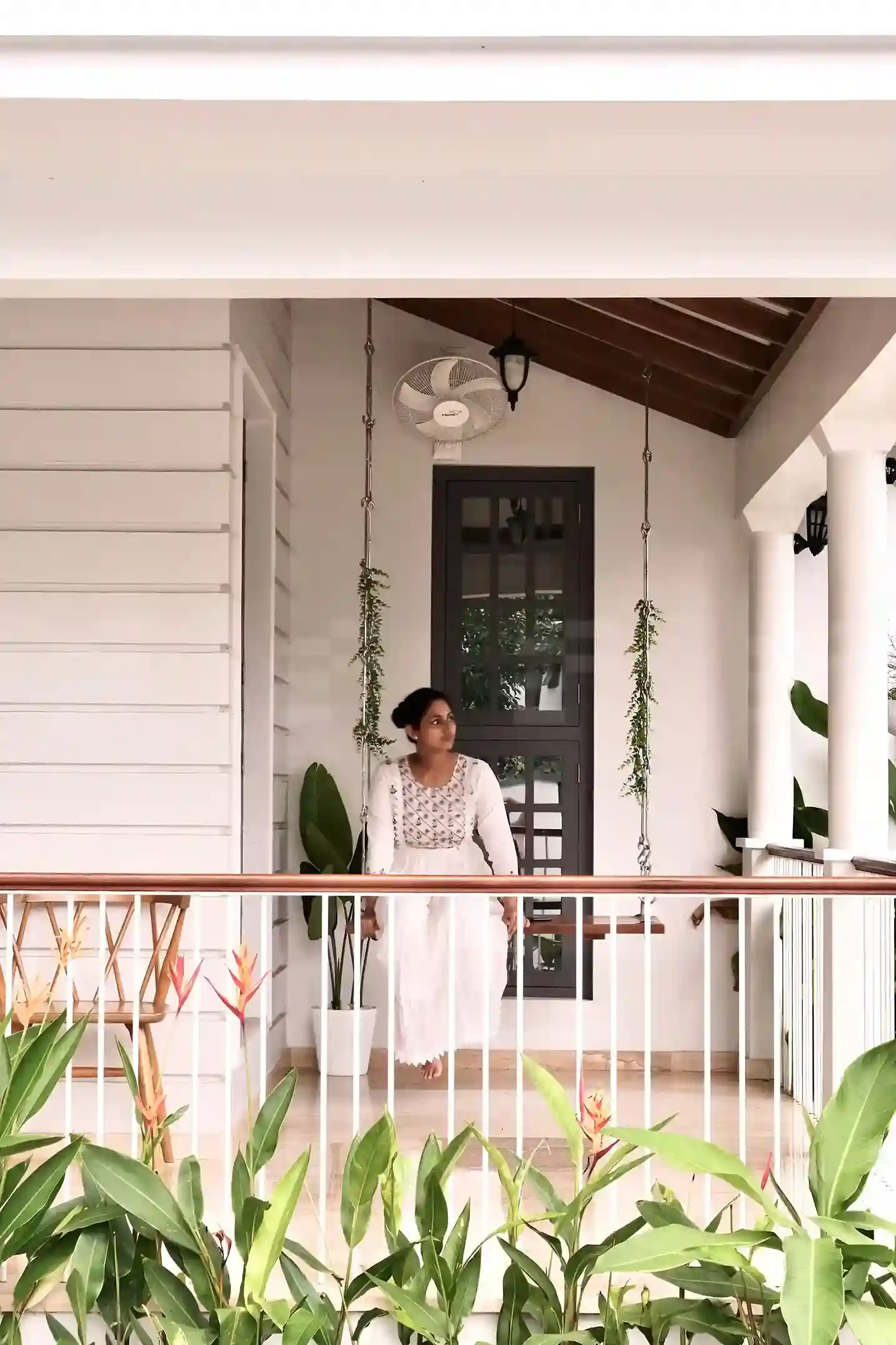olonial style sit-out with graceful colonnades, lush plants, and a girl relaxing on a wooden swing in a serene setting.