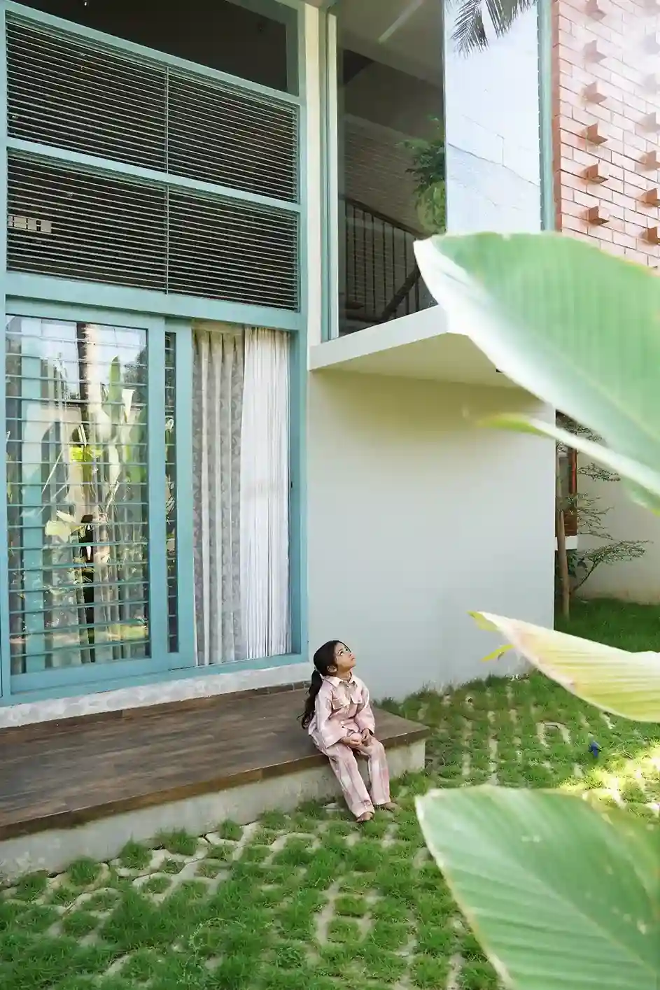 Contemporary house design Kerala – child seated on wooden deck with sliding windows and patterned grass paving.