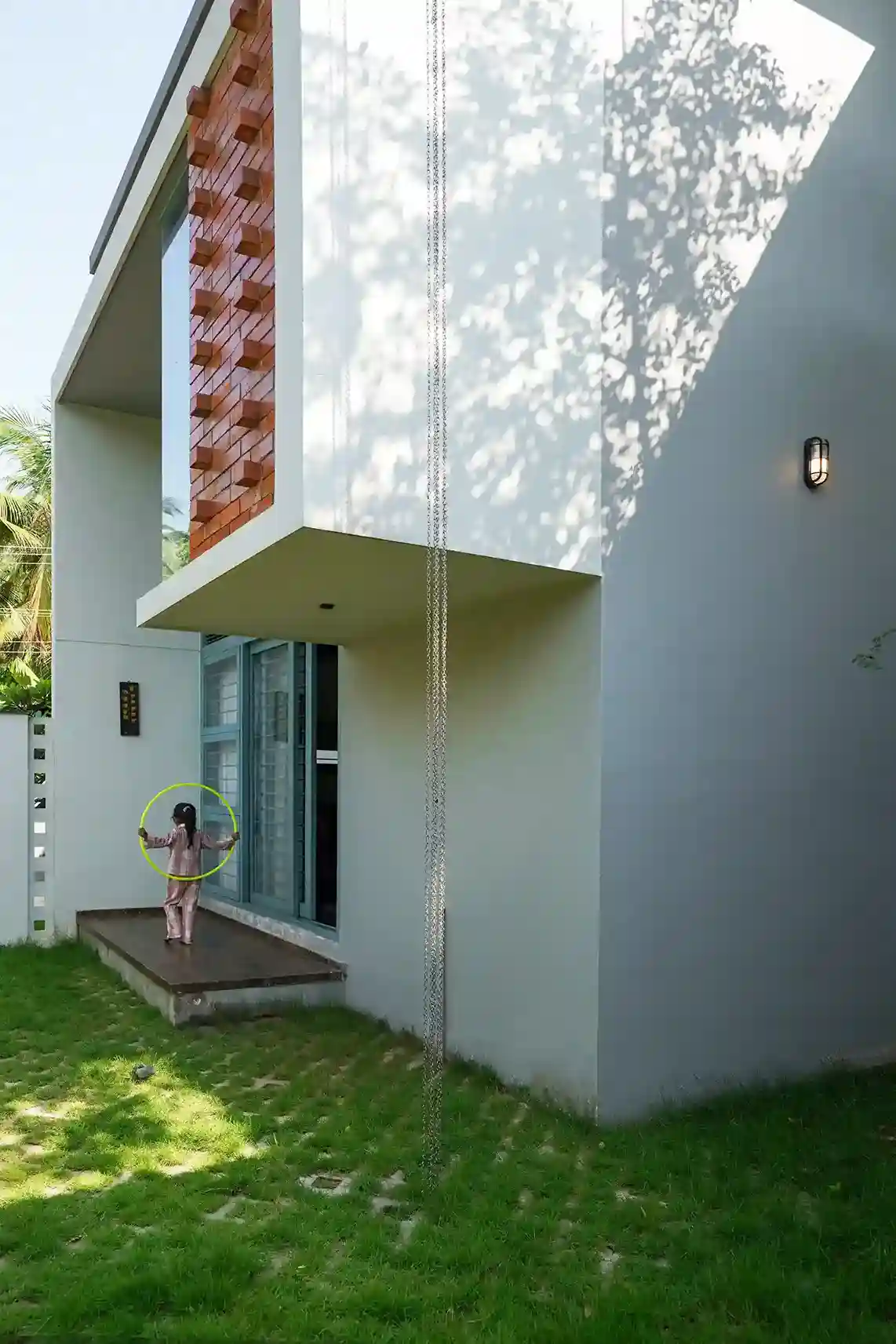Contemporary house design Kerala – child playing with hula hoop on wooden deck with brick jaali and rain chain.