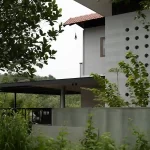 Side view of a tropical modern house design in Vettukad, Pulikkal, Kerala, featuring a concrete jali wall, terracota tile roof, and lush green surroundings.