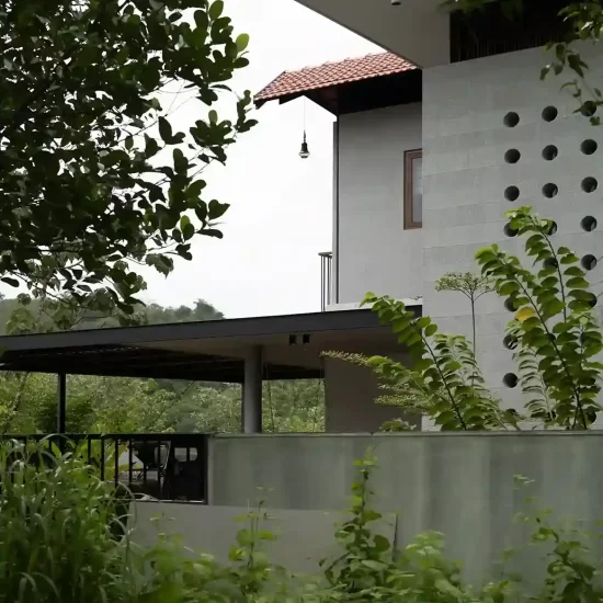 Side view of a tropical modern house design in Vettukad, Pulikkal, Kerala, featuring a concrete jali wall, terracota tile roof, and lush green surroundings.