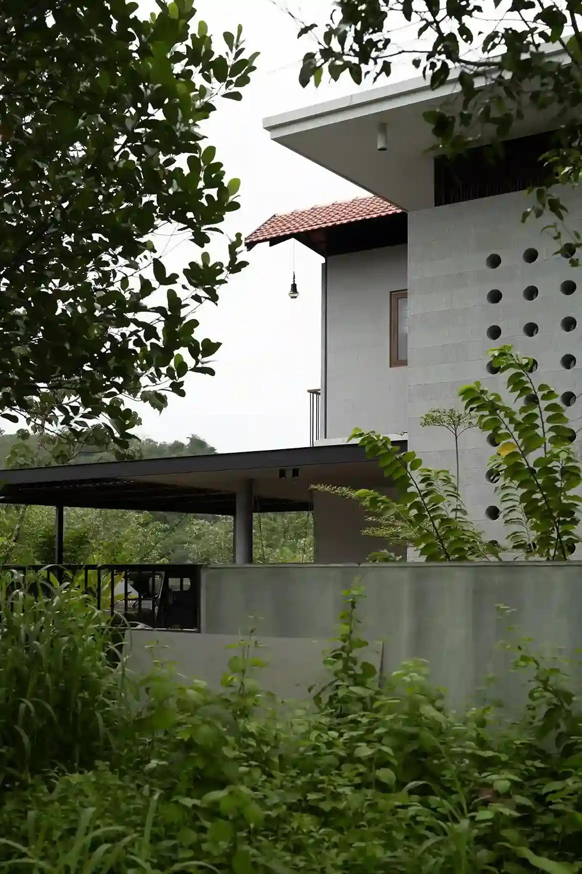 Side view of a tropical modern house design in Vettukad, Pulikkal, Kerala, featuring a concrete jali wall, terracota tile roof, and lush green surroundings.